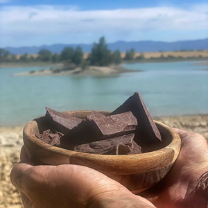 Peut inclure: Un bol en bois rempli de morceaux de chocolat brun foncé, tenu dans les mains d'une personne. Le bol est net, tandis que l'arrière-plan est flou et montre une étendue d'eau et un paysage lointain.