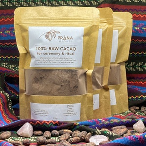 Peut inclure: Deux sacs en papier brun remplis de poudre de cacao brun foncé. Les sacs sont étiquetés "PRANA plants 100% RAW CACAO for ceremony & ritual". Les sacs sont posés sur un tissu à motifs colorés avec des fèves de cacao et des cristaux roses.