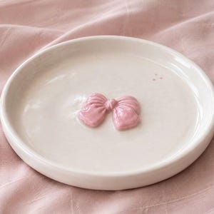 Pu&ograve; includere: Un piattino rotondo in ceramica color bianco sporco con un fiocco rosa chiaro lucido in rilievo al centro. L'oggetto presenta una finitura smaltata liscia e un bordo circolare semplice, offrendo una superficie d'appoggio pulita e funzionale.