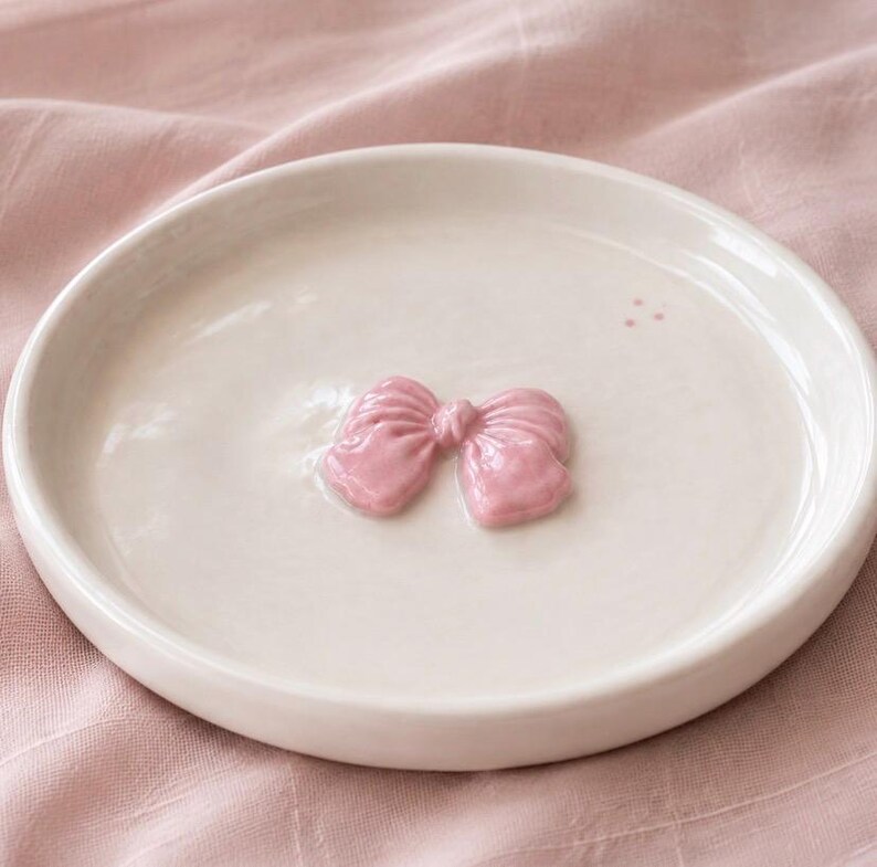 Pu&ograve; includere: Un piattino rotondo in ceramica color bianco sporco con un fiocco rosa chiaro lucido in rilievo al centro. L'oggetto presenta una finitura smaltata liscia e un bordo circolare semplice, offrendo una superficie d'appoggio pulita e funzionale.