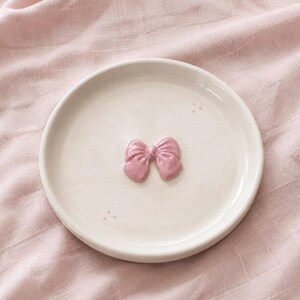 Pu&ograve; includere: Un piattino rotondo in ceramica color bianco sporco con un fiocco rosa lucido in rilievo al centro. Il piattino presenta due piccoli e sottili gruppi di tre minuscoli puntini rosa posizionati vicino ai bordi superiore destro e inferiore sinistro.