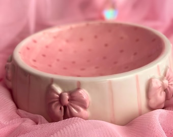 May include: A round ceramic bowl with a light pink interior featuring small darker pink polka dots. The exterior is off-white with vertical light pink stripes and three-dimensional pink bows attached around the outer rim of the bowl.
