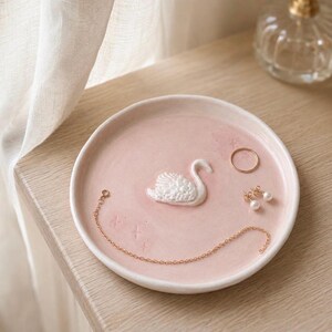 Pu&ograve; includere: Un piattino rotondo in ceramica rosa pallido con una figura di cigno bianco in rilievo al centro. Il piatto contiene una sottile catenina dorata, un semplice anello dorato e un paio di piccoli orecchini dorati con perle, oltre a motivi a stella.