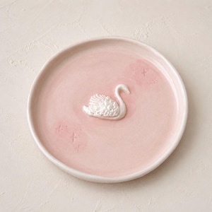 Pu&ograve; includere: Un piattino rotondo in ceramica rosa chiaro con una figura di cigno bianco in rilievo al centro. La superficie del piatto &egrave; decorata con piccole e sottili incisioni a forma di stella sparse attorno al cigno.