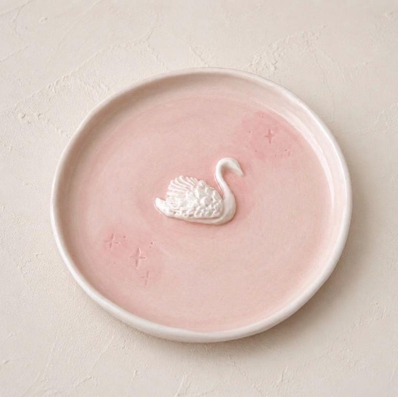 Pu&ograve; includere: Un piattino rotondo in ceramica rosa chiaro con una figura di cigno bianco in rilievo al centro. La superficie del piatto &egrave; decorata con piccole e sottili incisioni a forma di stella sparse attorno al cigno.