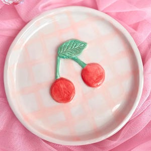 May include: A round white ceramic trinket dish featuring a hand-painted light pink checkered pattern across the surface. A raised, three-dimensional sculpted cherry motif with two bright red cherries and a single textured green leaf sits in the center.