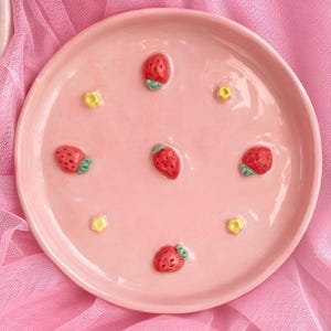 May include: A round, light pink ceramic plate featuring a glossy finish. The surface is decorated with four small, raised red strawberry motifs with green leaves and four tiny, raised yellow flower accents arranged in a circular pattern.