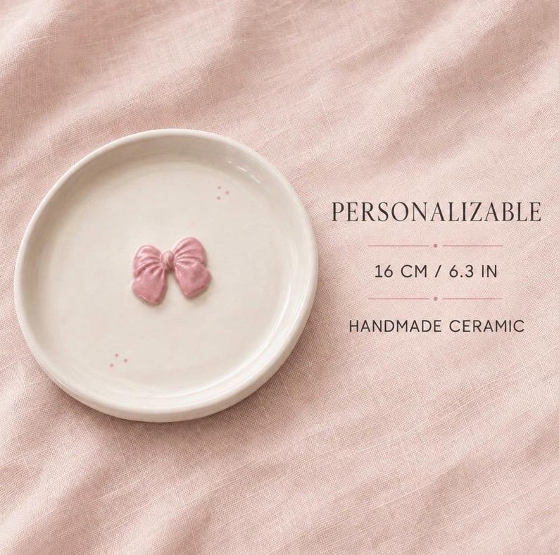 Pu&ograve; includere: Un piattino rotondo in ceramica color bianco sporco con un piccolo fiocco rosa in rilievo al centro e tre minuscoli punti vicino al bordo. Il testo riporta PERSONALIZABLE, 16 CM / 6.3 IN e HANDMADE CERAMIC. L'oggetto ha una finitura liscia e lucida.