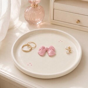Pu&ograve; includere: Un piattino rotondo in ceramica color bianco sporco con una piccola scultura a forma di fiocco rosa lucido al centro, due anelli dorati e un paio di orecchini a perla bianchi. Il piatto &egrave; decorato con minuscoli e delicati puntini rosa.