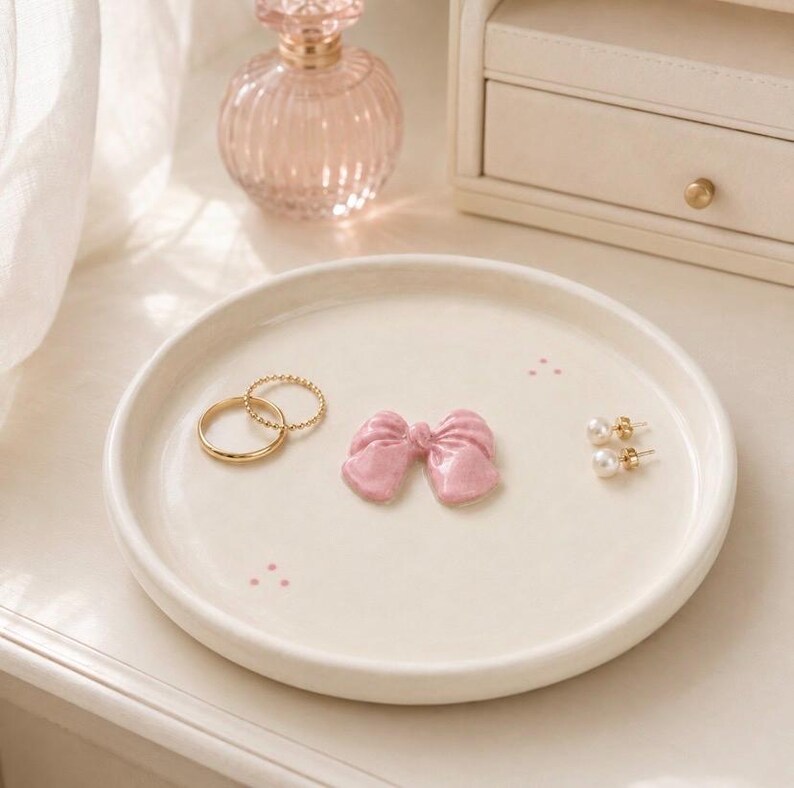 Pu&ograve; includere: Un piattino rotondo in ceramica color bianco sporco con una piccola scultura a forma di fiocco rosa lucido al centro, due anelli dorati e un paio di orecchini a perla bianchi. Il piatto &egrave; decorato con minuscoli e delicati puntini rosa.