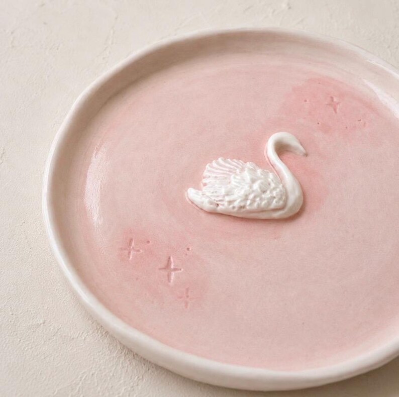 Pu&ograve; includere: Un piatto rotondo in ceramica rosa chiaro con una figura di cigno bianca tridimensionale in rilievo al centro. La superficie del piatto &egrave; decorata con piccole forme a stella in rilievo sparse attorno al cigno, creando un aspetto testurizzato.