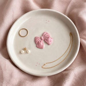 Pu&ograve; includere: Un piattino rotondo in ceramica color bianco sporco con un fiocco rosa scolpito in rilievo al centro. Il piatto contiene un sottile anello dorato, un paio di orecchini a perla e una delicata collana a catena dorata, con piccoli punti rosa.