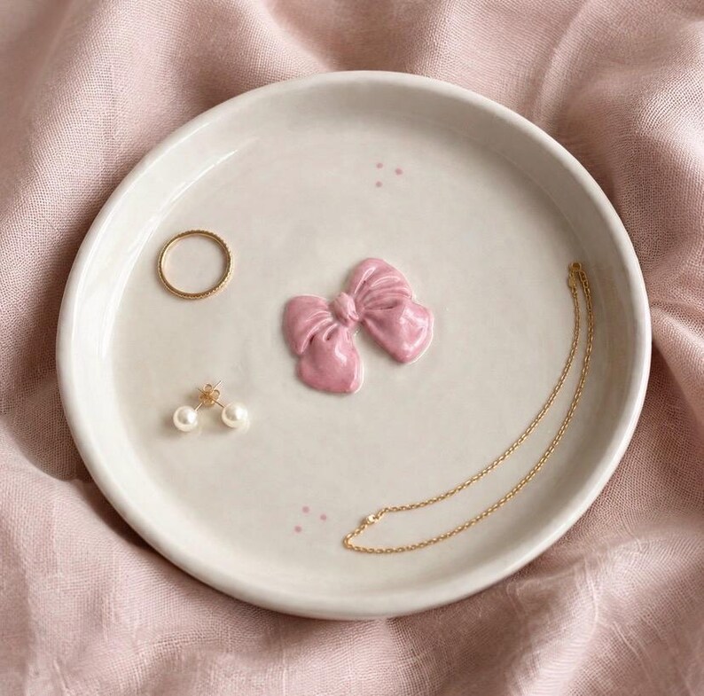 Pu&ograve; includere: Un piattino rotondo in ceramica color bianco sporco con un fiocco rosa scolpito in rilievo al centro. Il piatto contiene un sottile anello dorato, un paio di orecchini a perla e una delicata collana a catena dorata, con piccoli punti rosa.