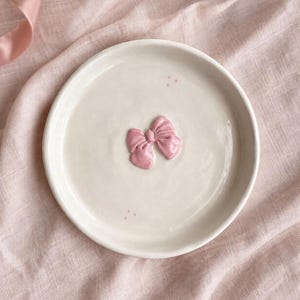 Pu&ograve; includere: Un piattino rotondo in ceramica color bianco sporco con un fiocco rosa scolpito in rilievo al centro. La superficie del piattino &egrave; liscia e lucida, con piccoli e delicati puntini rosa posizionati sopra e sotto la decorazione centrale.