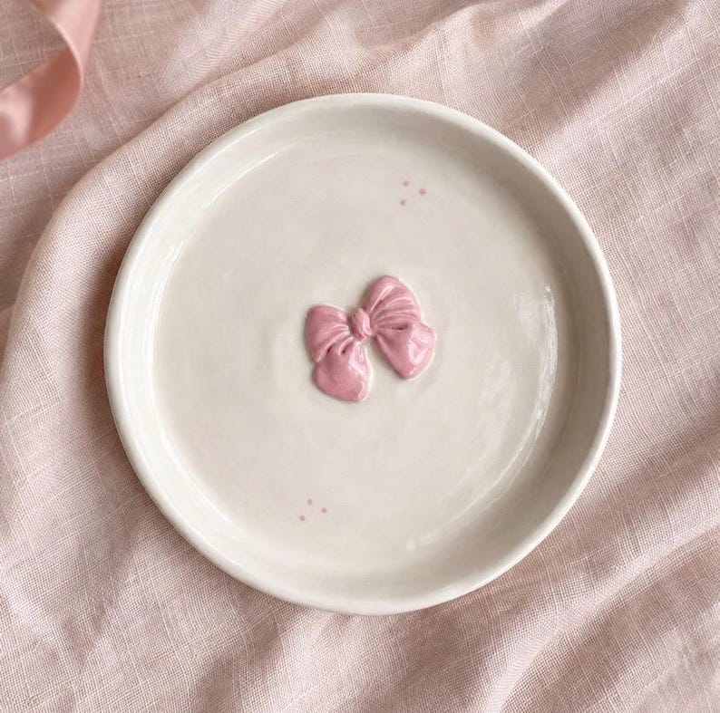 Pu&ograve; includere: Un piattino rotondo in ceramica color bianco sporco con un fiocco rosa scolpito in rilievo al centro. La superficie del piattino &egrave; liscia e lucida, con piccoli e delicati puntini rosa posizionati sopra e sotto la decorazione centrale.