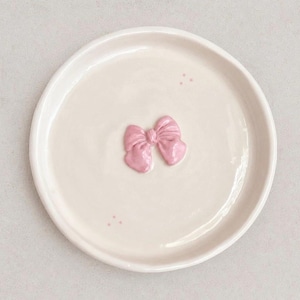 Pu&ograve; includere: Un piattino rotondo in ceramica color bianco sporco con un fiocco rosa tridimensionale in rilievo al centro. La superficie del piatto &egrave; liscia e lucida, accentuata da due piccoli gruppi di tre minuscoli puntini rosa vicino al bordo.