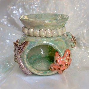 May include: A ceramic teal and green oil burner with a mermaid tail and starfish design. The burner has a pearl-like trim around the top.