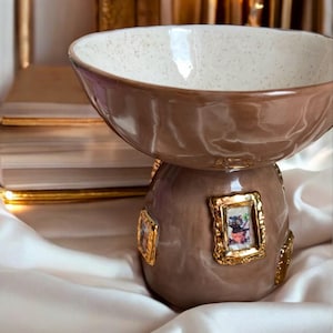 May include: A ceramic bowl with a wide, glossy brown rim and a speckled cream interior. The base is a rounded brown pedestal decorated with multiple small, ornate gold-colored rectangular frames, each containing a miniature colorful illustration.