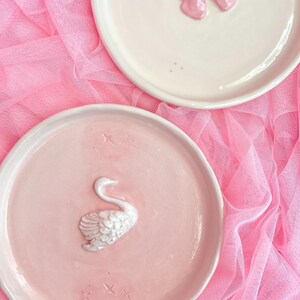 Pu&ograve; includere: Due piatti rotondi in ceramica. Il piatto superiore &egrave; bianco sporco con un fiocco rosa lucido in rilievo al centro e piccoli punti rosa. Il piatto inferiore &egrave; rosa chiaro con una figura di cigno bianca e testurizzata in rilievo al centro.