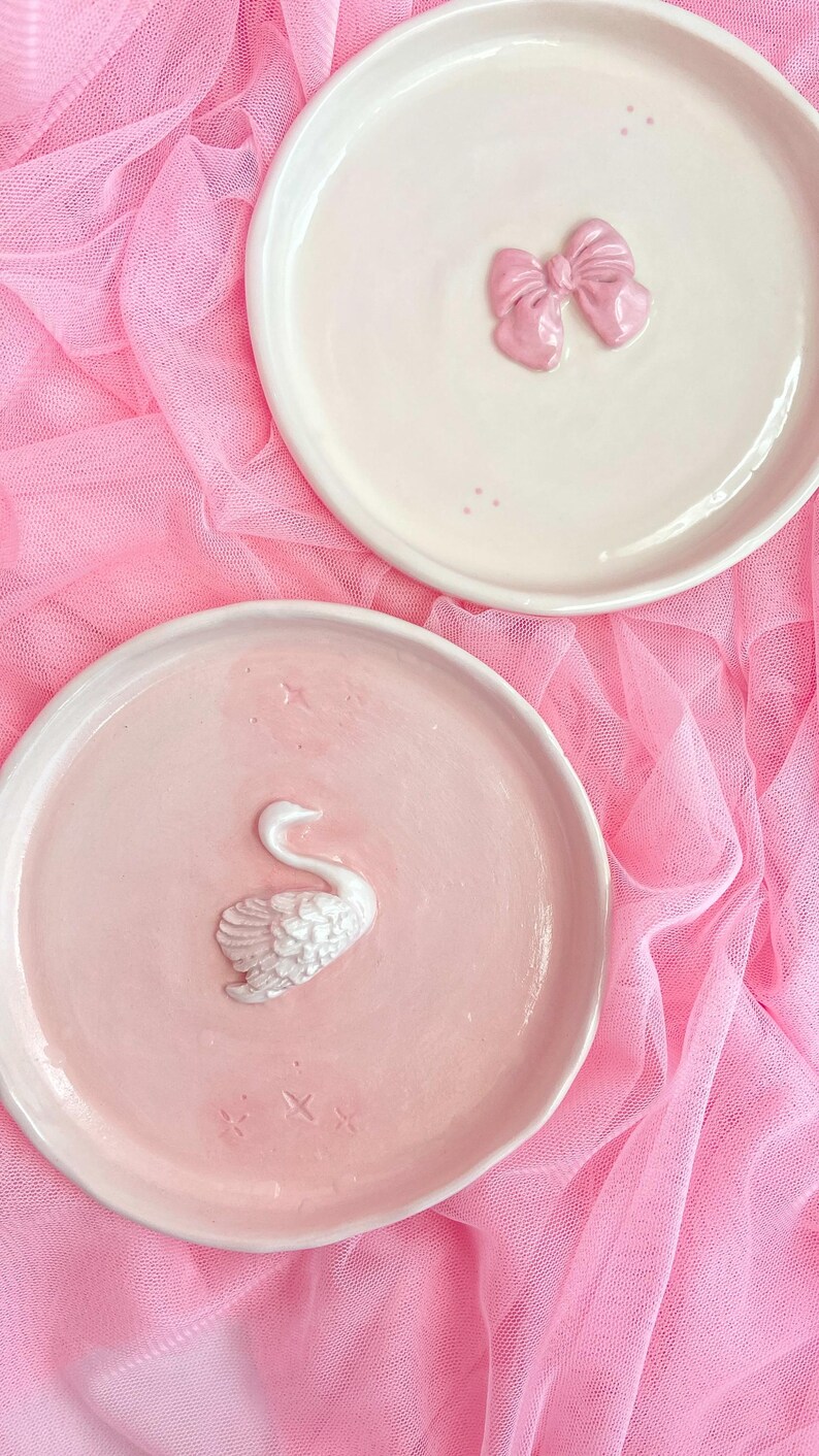 Pu&ograve; includere: Due piatti rotondi in ceramica. Il piatto superiore &egrave; bianco sporco con un fiocco rosa lucido in rilievo al centro e piccoli punti rosa. Il piatto inferiore &egrave; rosa chiaro con una figura di cigno bianca e testurizzata in rilievo al centro.