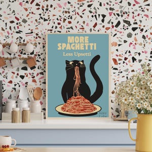 May include: A rectangular wall art print with a light blue background featuring a black cat eating a large pile of red spaghetti on a plate. The text reads MORE SPAGHETTI Less Upsetti in yellow and white letters. The artist name Kara Labs is visible.