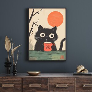 May include: A framed art print featuring a black cat holding an orange mug with a design. The cat is set against a backdrop of a rising orange sun and a light beige sky. The art has a Japanese aesthetic.