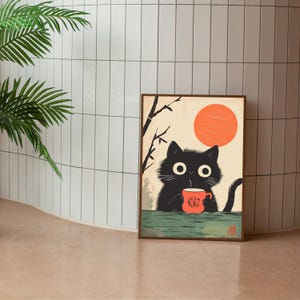 May include: A framed art print featuring a black cat holding a red mug with a logo, set against a backdrop of a rising orange sun and a beige sky. The cat is depicted in a minimalist style, with a green table.