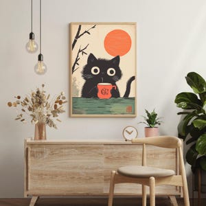 May include: A framed art print featuring a black cat holding a red mug with a design. The cat has large, round eyes and is set against a backdrop of a rising orange sun and stylized branches. The print has a wooden frame.