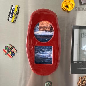 May include: A red fridge magnet with a rectangular shape and two cut-out windows showing a coastal scene with ocean waves. The magnet is surrounded by other decorative magnets.