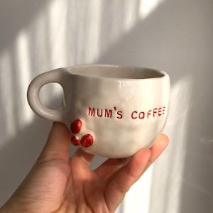 May include: White ceramic coffee mug with a red inscription that reads "MUM'S COFFEE". The mug has three red coffee bean accents.