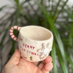 May include: A white ceramic mug with a red and white candy cane handle. The mug has a green and red Christmas tree design and the words "Merry Christmas" in red lettering. A white snowflake is attached to the side of the mug.
