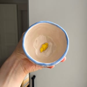 May include: A small, light blue ceramic bowl with a white interior and a yellow design in the centre. The bowl is being held by a hand.