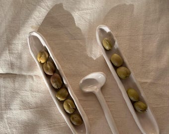 May include: A white ceramic serving spoon and two long, narrow ceramic dishes, each filled with green beans. The spoon has a rounded bowl and a long handle. The dishes are oval-shaped. The beans are a light green color.