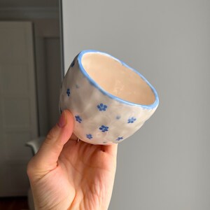 Puede incluir: Un peque&ntilde;o cuenco de cer&aacute;mica blanca con un borde azul y un patr&oacute;n floral azul. El cuenco est&aacute; ligeramente deformado y tiene una superficie texturizada.