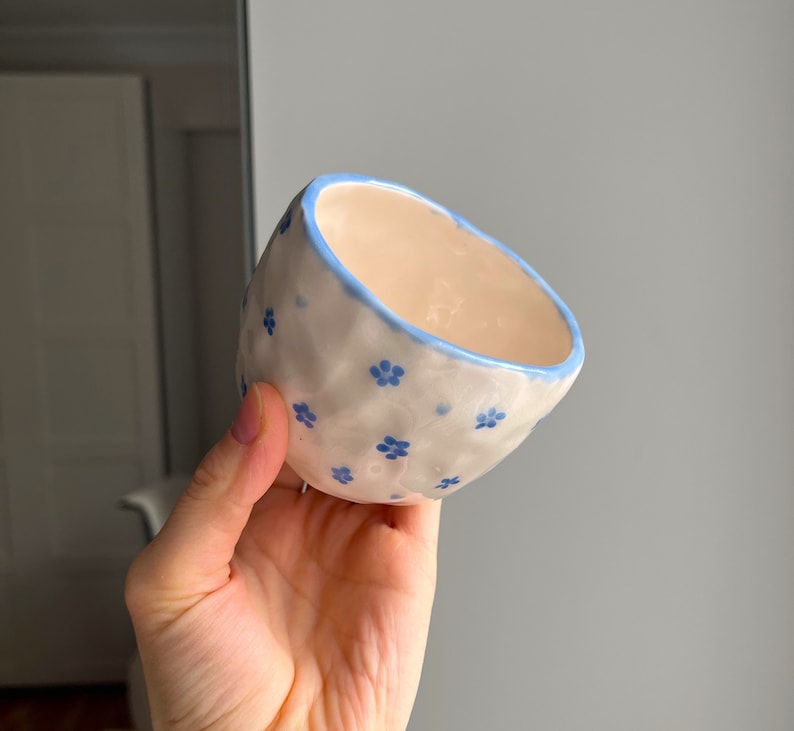 Puede incluir: Un peque&ntilde;o cuenco de cer&aacute;mica blanca con un borde azul y un patr&oacute;n floral azul. El cuenco est&aacute; ligeramente deformado y tiene una superficie texturizada.