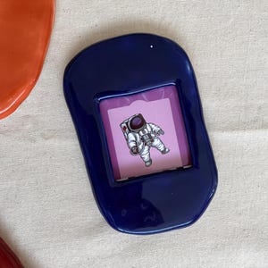 May include: A dark blue ceramic picture frame with a rounded shape. The frame holds a small image of an astronaut in a spacesuit against a pink background. The frame is placed on a neutral-colored surface.