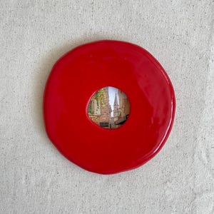 May include: A circular, glossy red ceramic object with an irregular, organic edge. The center features a circular cutout revealing a photographic image of a stone building with a tall spire, green foliage, and a lantern against a clear sky.