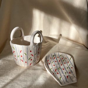 May include: White ceramic basket with two handles and a matching spoon rest. Both pieces are decorated with vertical blue lines and red and green chili pepper designs. The basket and spoon rest are on a white cloth.