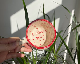 May include: A white ceramic cup with a red rim and red hearts. The inside of the cup says "I love you" in red lettering.