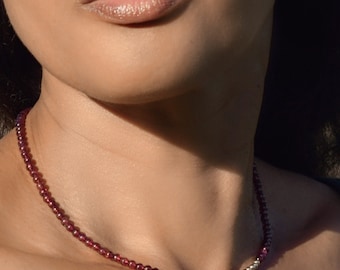 Op de afbeelding: Een delicate ketting gemaakt van kleine, donkerrode granaat kralen. De ketting wordt gedragen door een persoon met een lichte huidskleur.