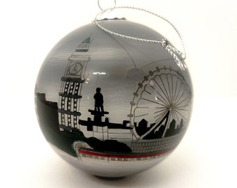 May include: A spherical glass Christmas ornament with a silver finish, featuring a hand-painted black silhouette of the London skyline, including Big Ben and the London Eye, with a silver hanging string attached to the top cap.