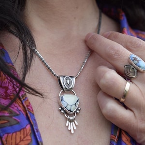 Könnte beinhalten: Eine silberne Halskette mit einem Anhänger mit einem weißen und schwarzen dreieckigen Stein. Die Halskette hat eine strukturierte Kette und wird über einem bunten Oberteil mit Blumenmuster getragen. Ringe mit blauen Steinen und ein goldenes Band sind ebenfalls sichtbar.