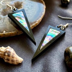 May include: A pair of dangle earrings featuring black triangular frames with iridescent abalone shell inlays. Each earring is suspended from a silver-toned metal hook, showcasing a geometric design with a vibrant, multi-colored pearlescent finish.