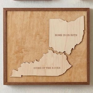 May include: Wooden wall art featuring the states of Ohio and Kentucky, with the text "HOME IS ON BOTH SIDES OF THE RIVER" engraved on the wood. The artwork is framed in a dark brown wooden frame.