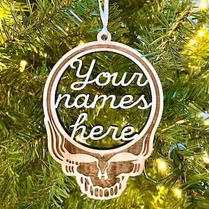 May include: Wooden Christmas ornament in the shape of a skull with the words "Your names here" cut out in a cursive font. The ornament is hanging from a white ribbon against a backdrop of a Christmas tree.