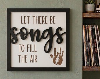 Può includere: Un cartello da parete quadrato con cornice nera e sfondo bianco. Il testo recita LET THERE BE in marrone, seguito dalla parola songs in grande corsivo nero e TO FILL THE AIR in marrone. È inclusa un'impronta di mano marrone con un fulmine.