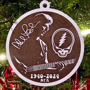 May include: A round, brown wooden ornament with a white outline. It features a silhouette of a person playing a guitar, a skull with a lightning bolt, and the text "1940-2024 NFA".