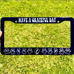 May include: Black license plate frame with the text "HAVE A GRATEFUL DAY" in white. The frame features dancing bears and a lightning bolt design. The frame is held against a green grassy background.