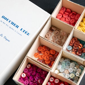 Peut inclure: Une boîte de boutons assortis de différentes couleurs et tailles, notamment rouge, orange, marron, jaune, vert et bleu. La boîte est étiquetée "JOS. BOUCHER LTÉE" et "Boutons La Vogue".