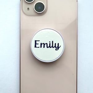 May include: A white phone case with a round, white pop socket that says "Emily" in blue lettering.